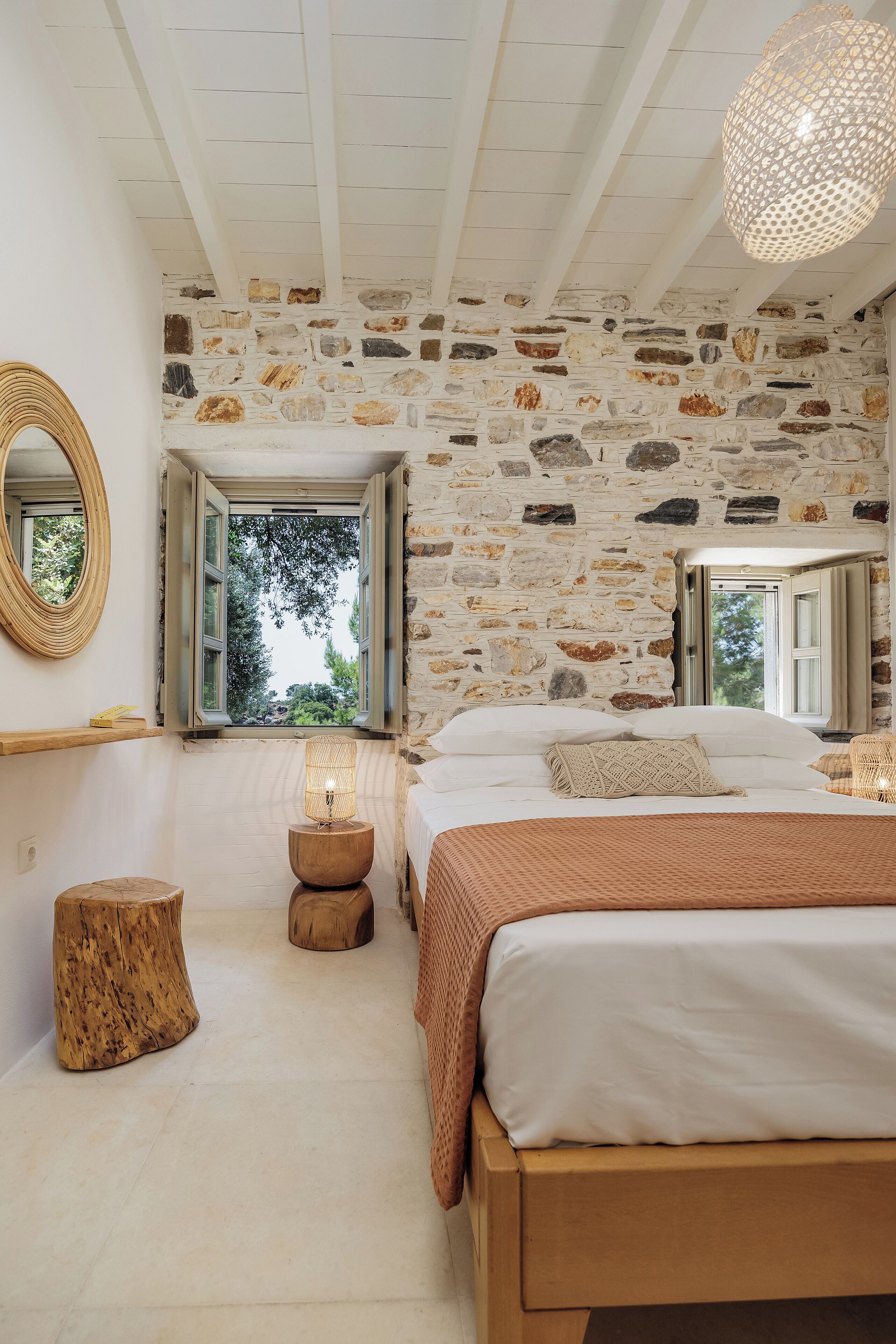 Une chambre avec un mur en pierre, un lit doté d’une tête de lit en bois, un miroir rond et une fenêtre aux volets ouverts.
