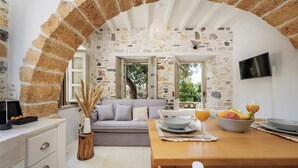 A cozy room with a stone wall, a wooden table set with fruit and glasses, a grey sofa, and a TV mounted on the wall. - Zos Suites (Naxos)