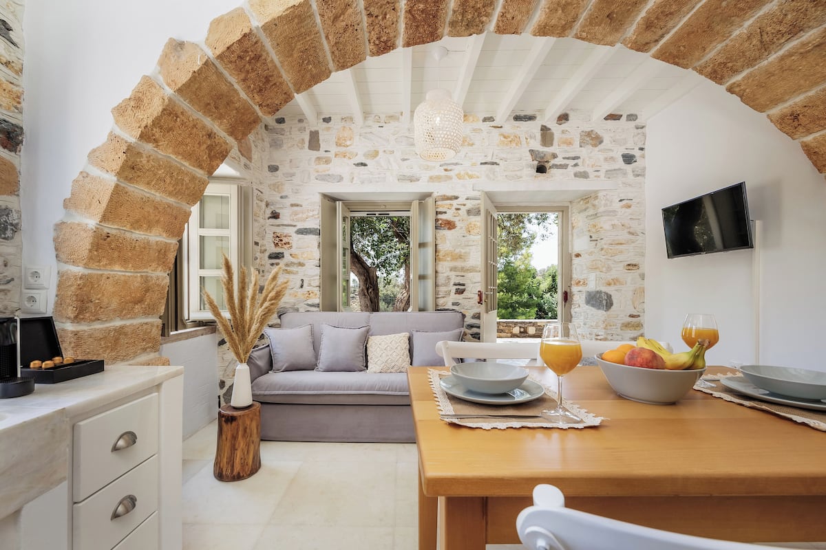 Um quarto aconchegante com parede de pedra, uma mesa de madeira posta com frutas e copos, um sofá cinza e uma TV fixada na parede.