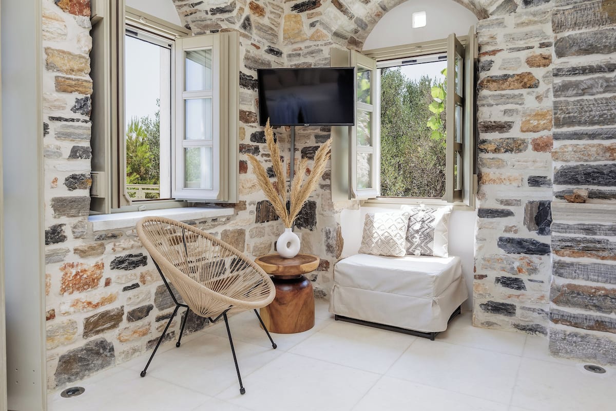 Quarto com paredes de pedra, uma televisão, uma cadeira de vime, uma pequena mesa com um vaso e um sofá.