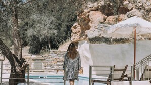 A poolside area with a stone wall, lounge chairs, and a person walking. - Zos Suites (Naxos)