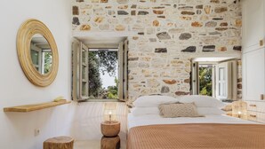 A bedroom with a stone wall, a round mirror, a bedside table with a lamp, a bed with a brown blanket, and a wooden stool. - Zos Suites (Naxos)
