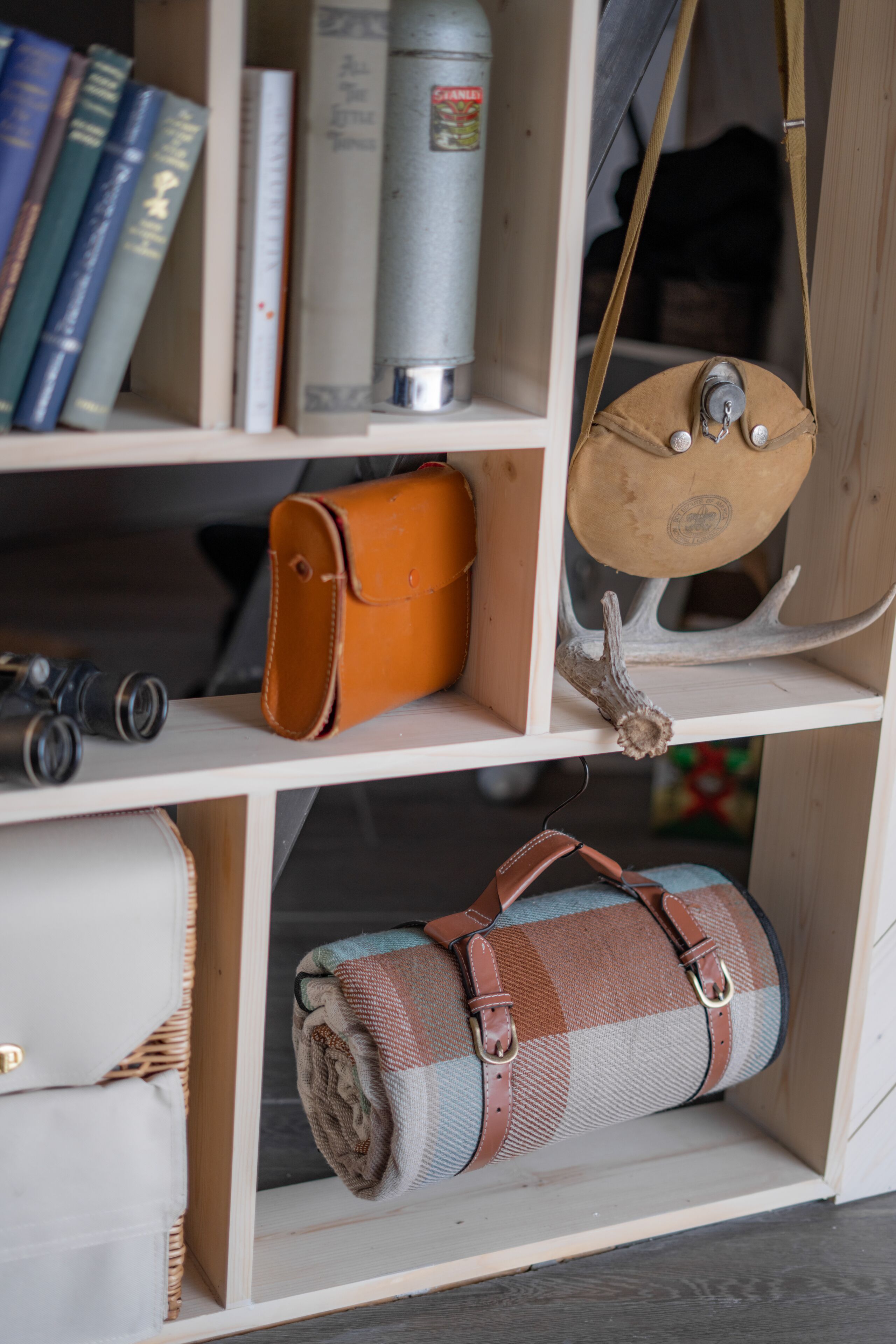 Uma prateleira de madeira com livros, uma bolsa de couro, um cobertor enrolado, binóculos e uma bombona de gás.