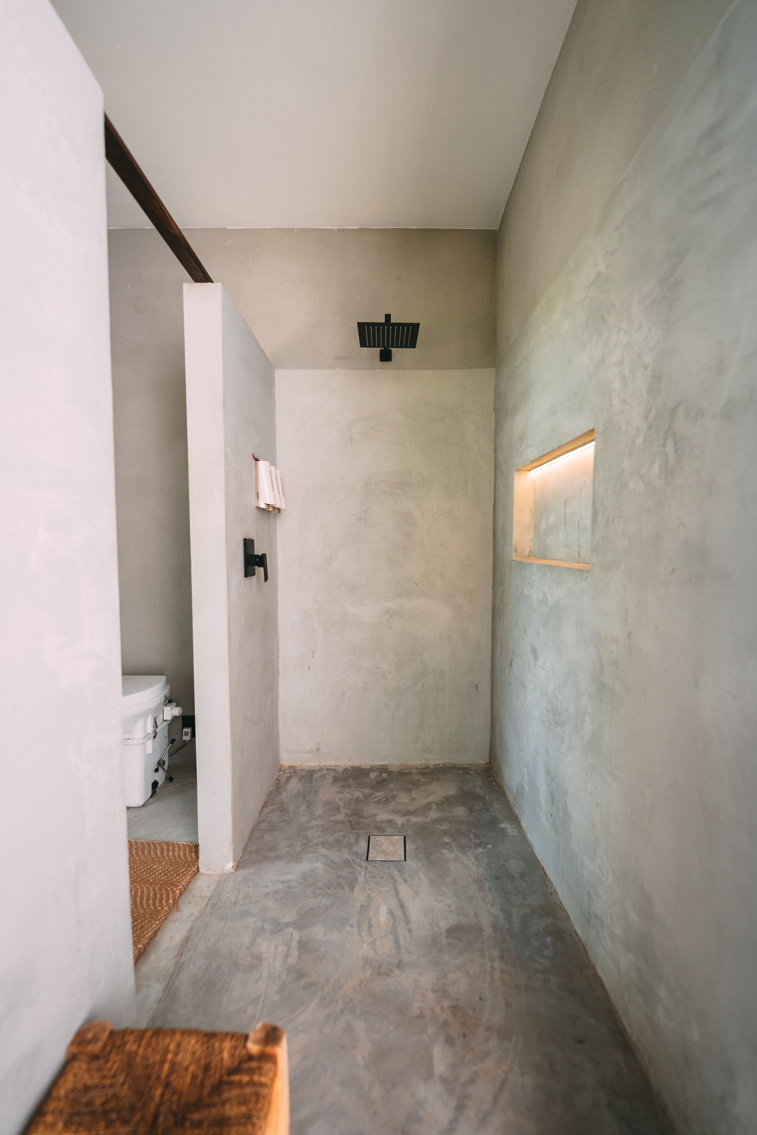 Un pasillo estrecho con suelo de hormigón, una pared blanca a un lado y una pared texturizada al otro. Se ve una pequeña ventana con una lámpara en la pared texturizada.