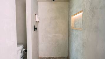 Un pasillo estrecho con suelo de hormigón, una pared blanca a un lado y una pared texturizada al otro. Se ve una pequeña ventana con una lámpara en la pared texturizada.