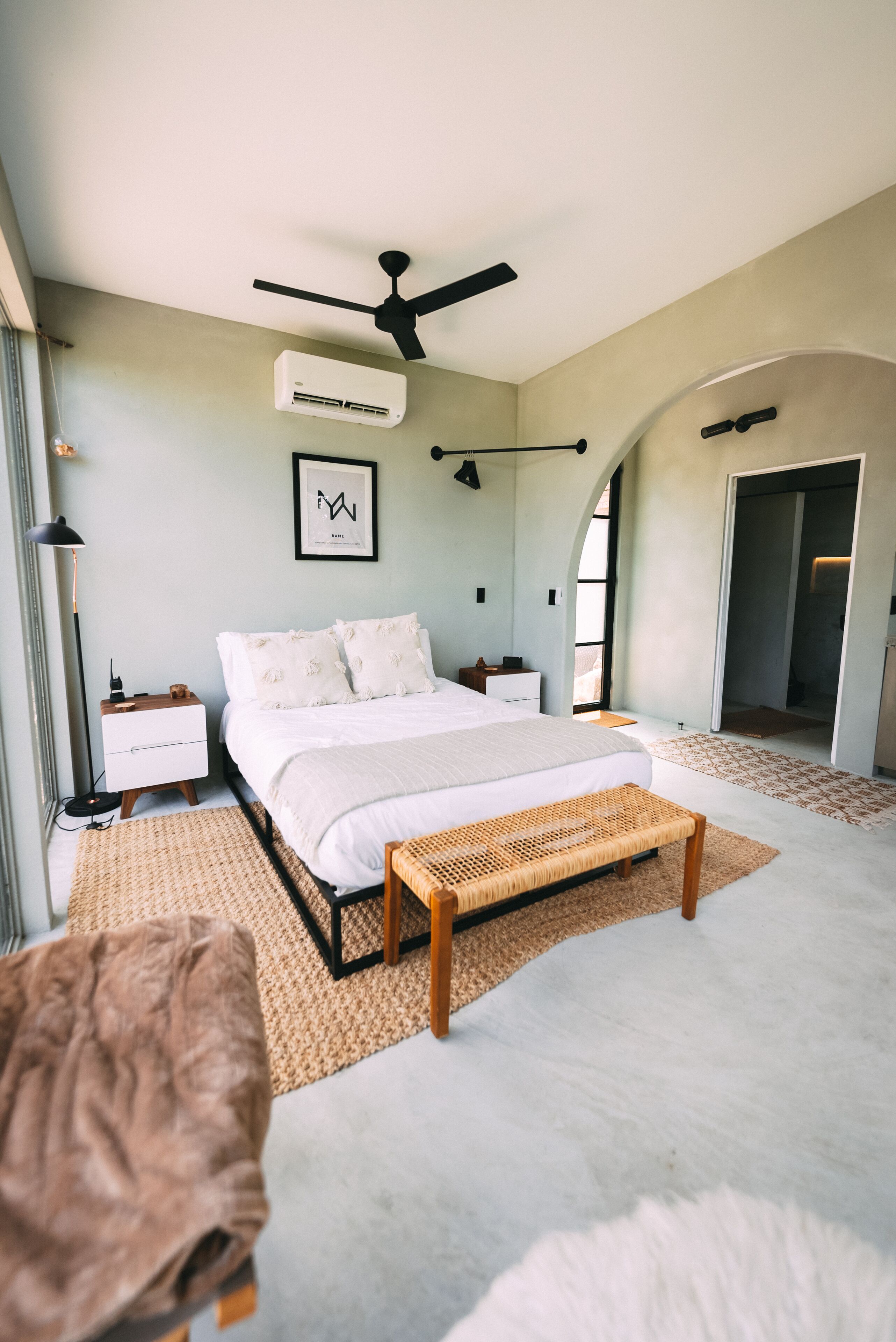 A modern bedroom with a large bed, a ceiling fan, an air conditioning unit, a bedside table, and a large window.