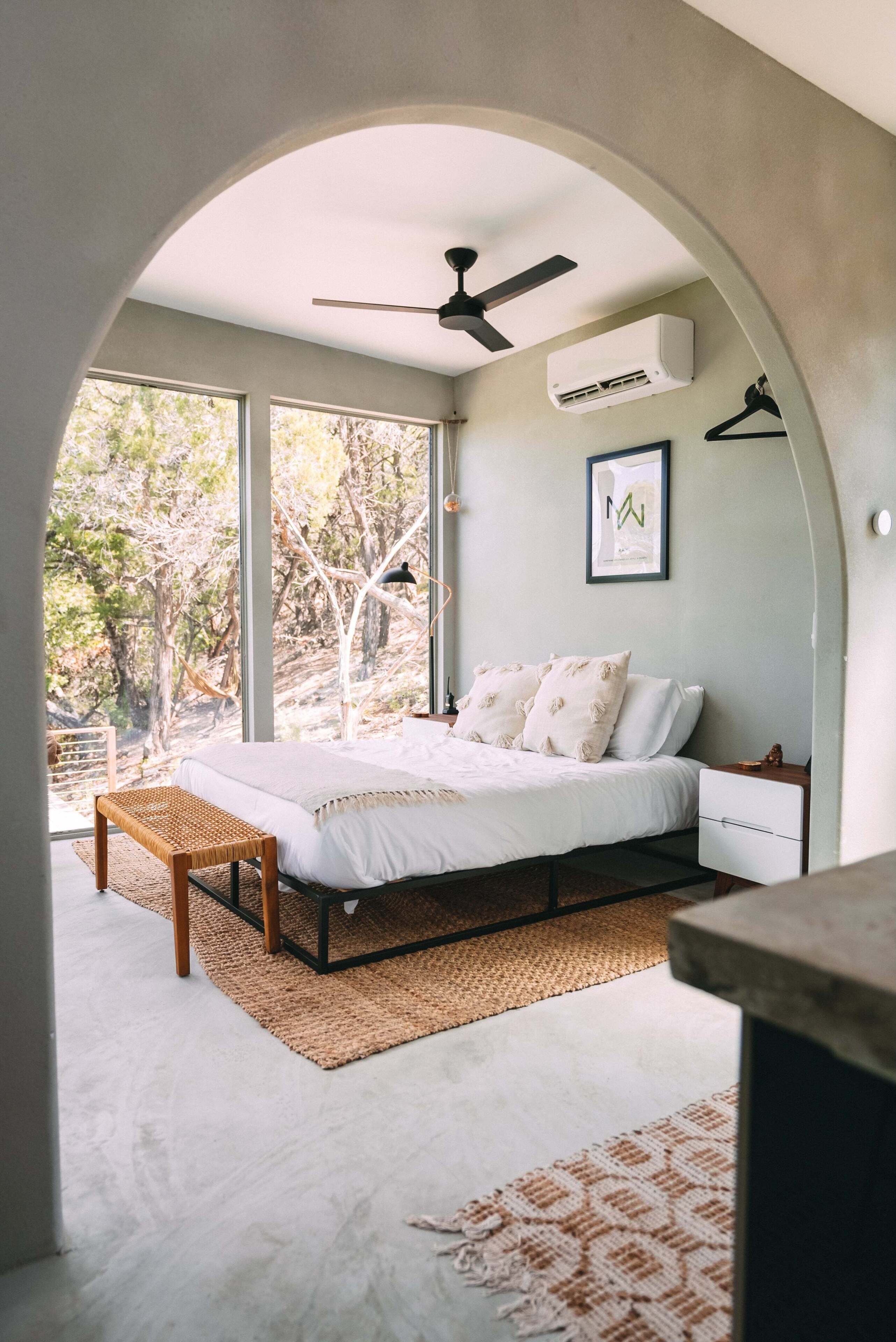 A bedroom with a bed, a ceiling fan, and a large window overlooking trees.