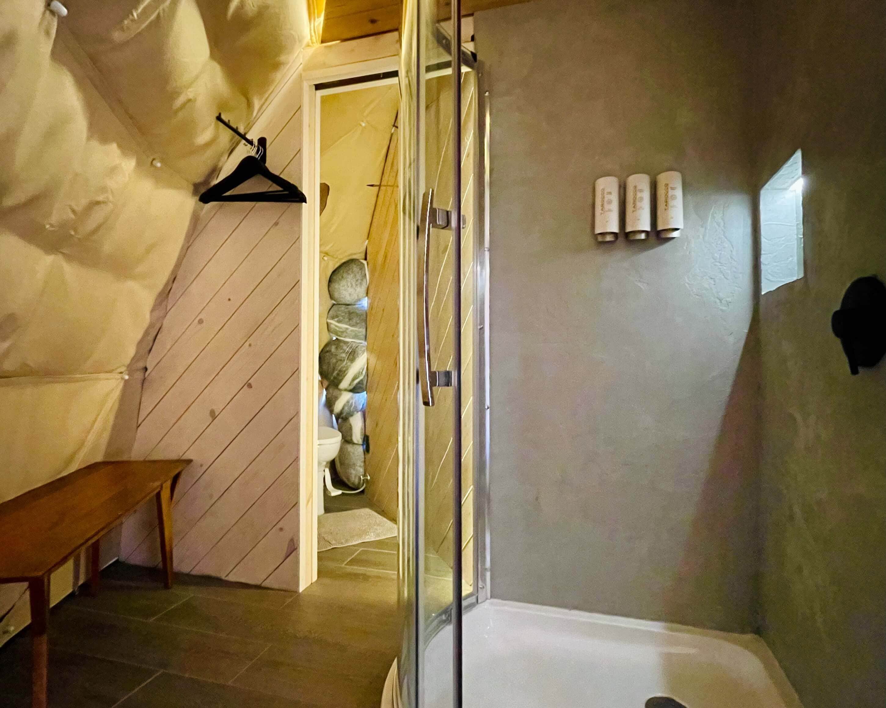 A bathroom with a glass shower enclosure, a wooden bench, and a stone wall.