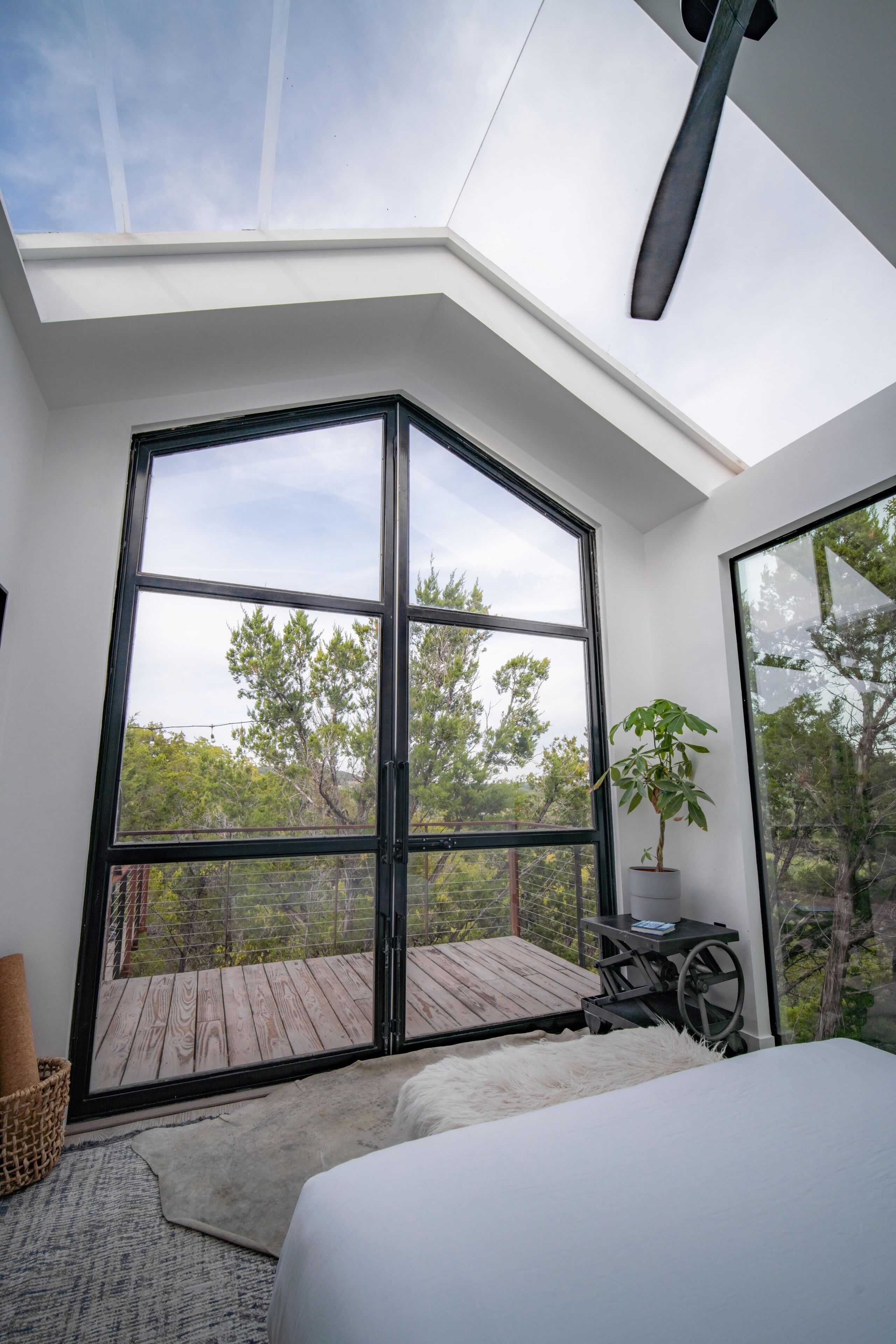 A bedroom with a large window, a bed, a small table, and a potted plant.