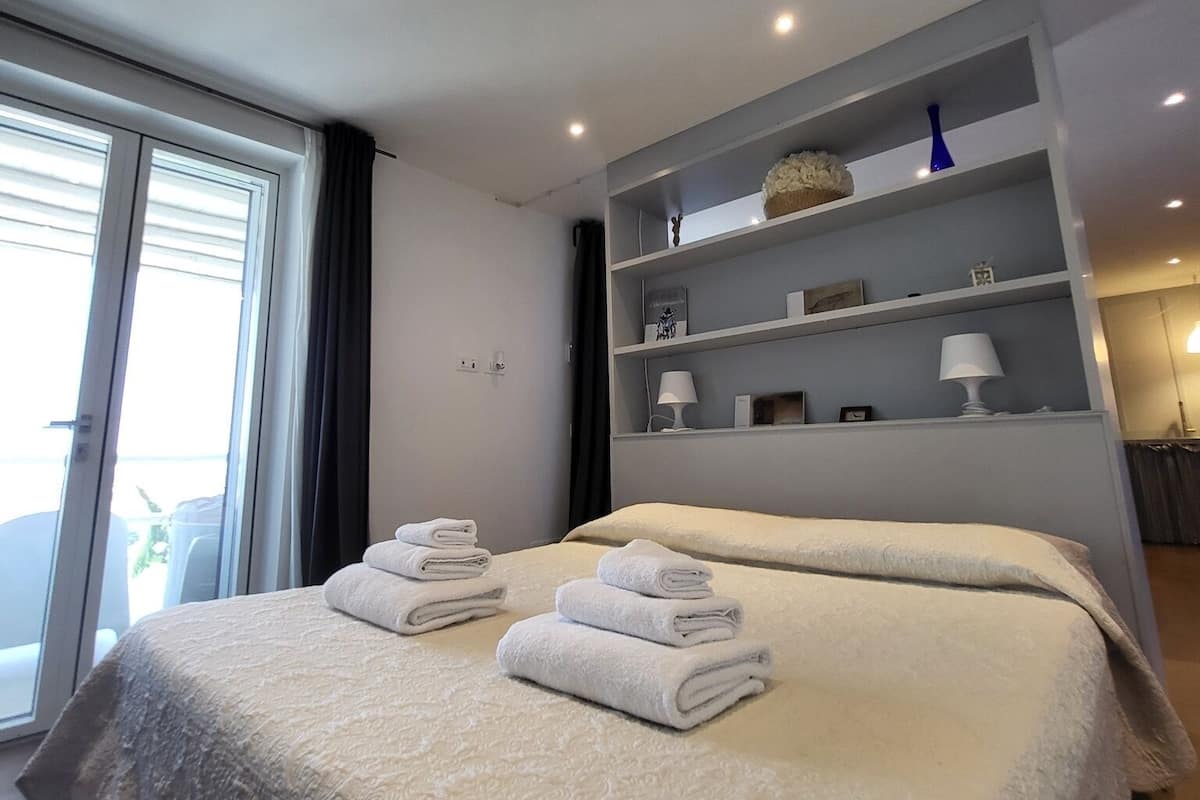 Quarto de hotel moderno com uma cama grande, uma estante embutida com objetos decorativos e vista para o exterior através de uma porta de correr.