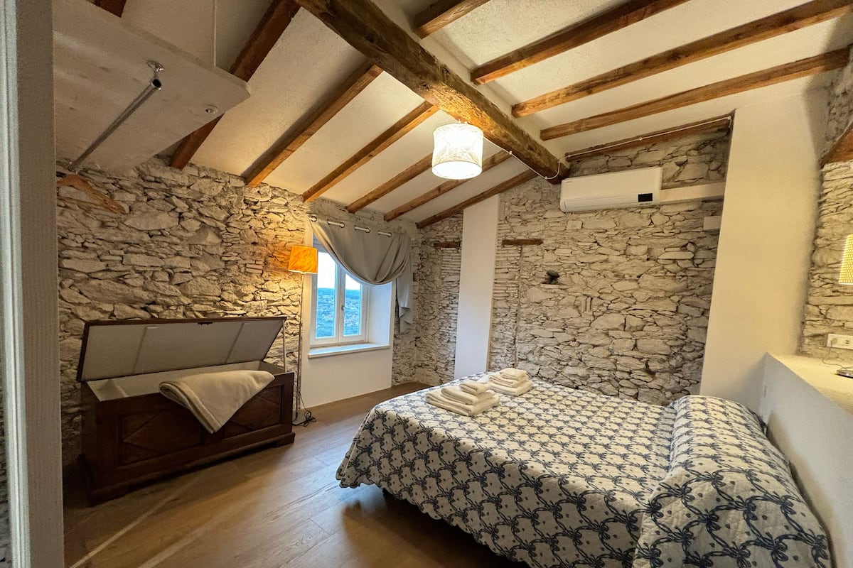 Quarto com parede de pedra, vigas de madeira, cama com roupa de cama estampada, mesa de cabeceira e janela com cortinas.