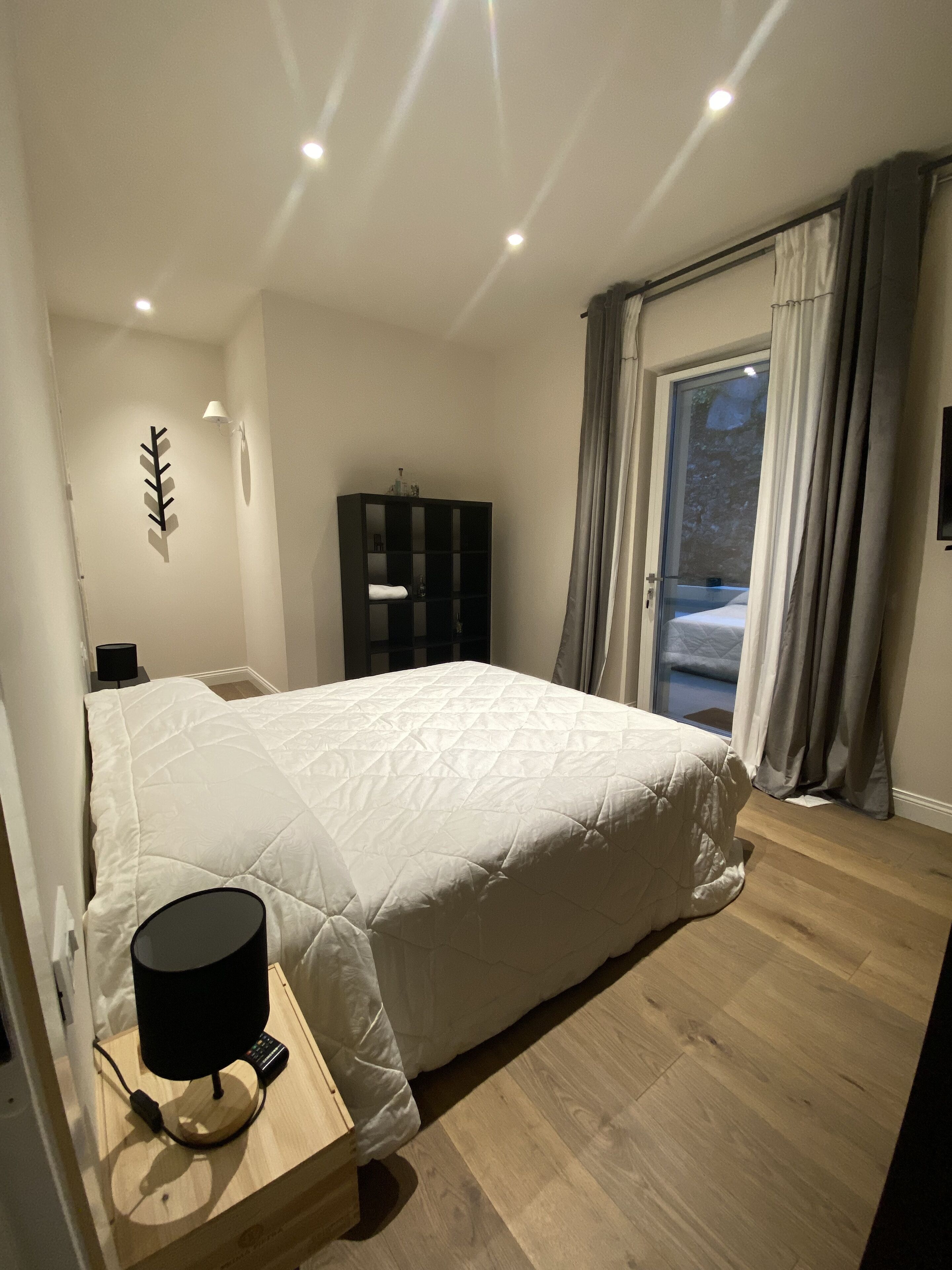 Ein Schlafzimmer mit einem großen Bett, einem Nachttisch mit Lampe, einem Kleiderschrank und Blick ins Freie durch eine Glastür.