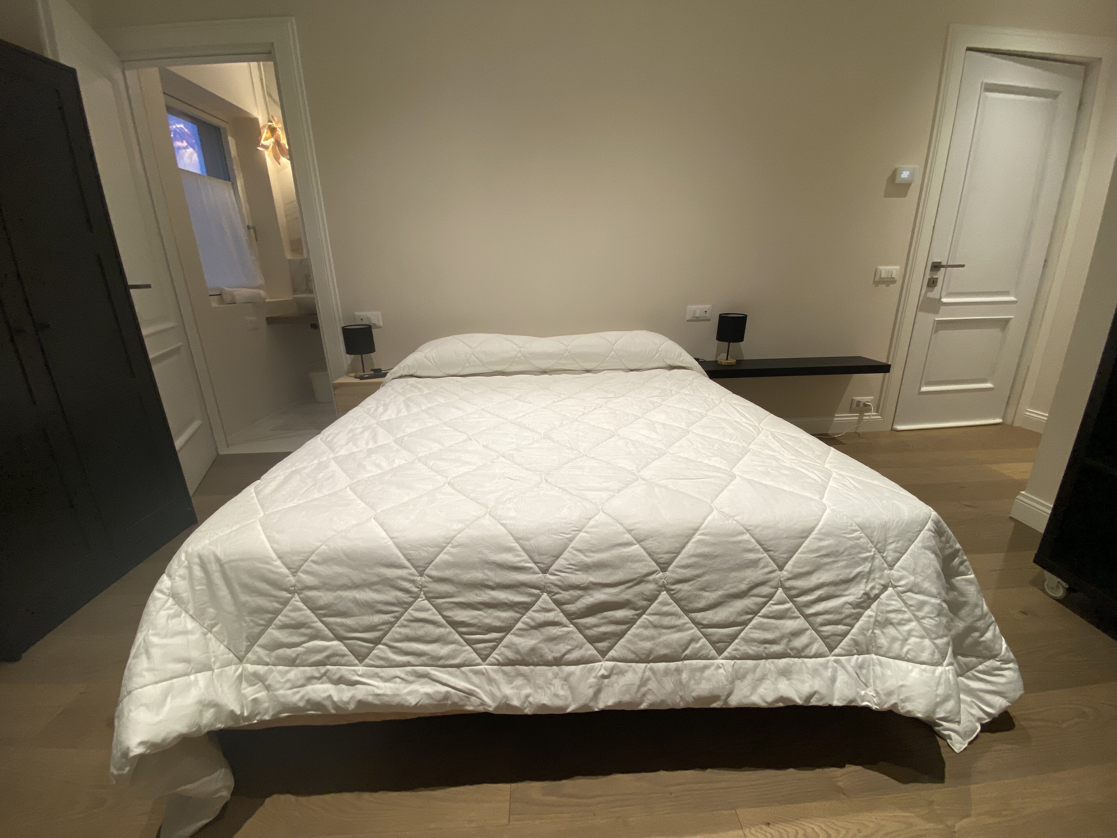 Un lit bien fait, recouvert d’une couette blanche, se trouve dans une pièce dont le sol est en bois et les murs sont peints en blanc. On y aperçoit une petite table de chevet avec une lampe, et une étagère au-dessus.