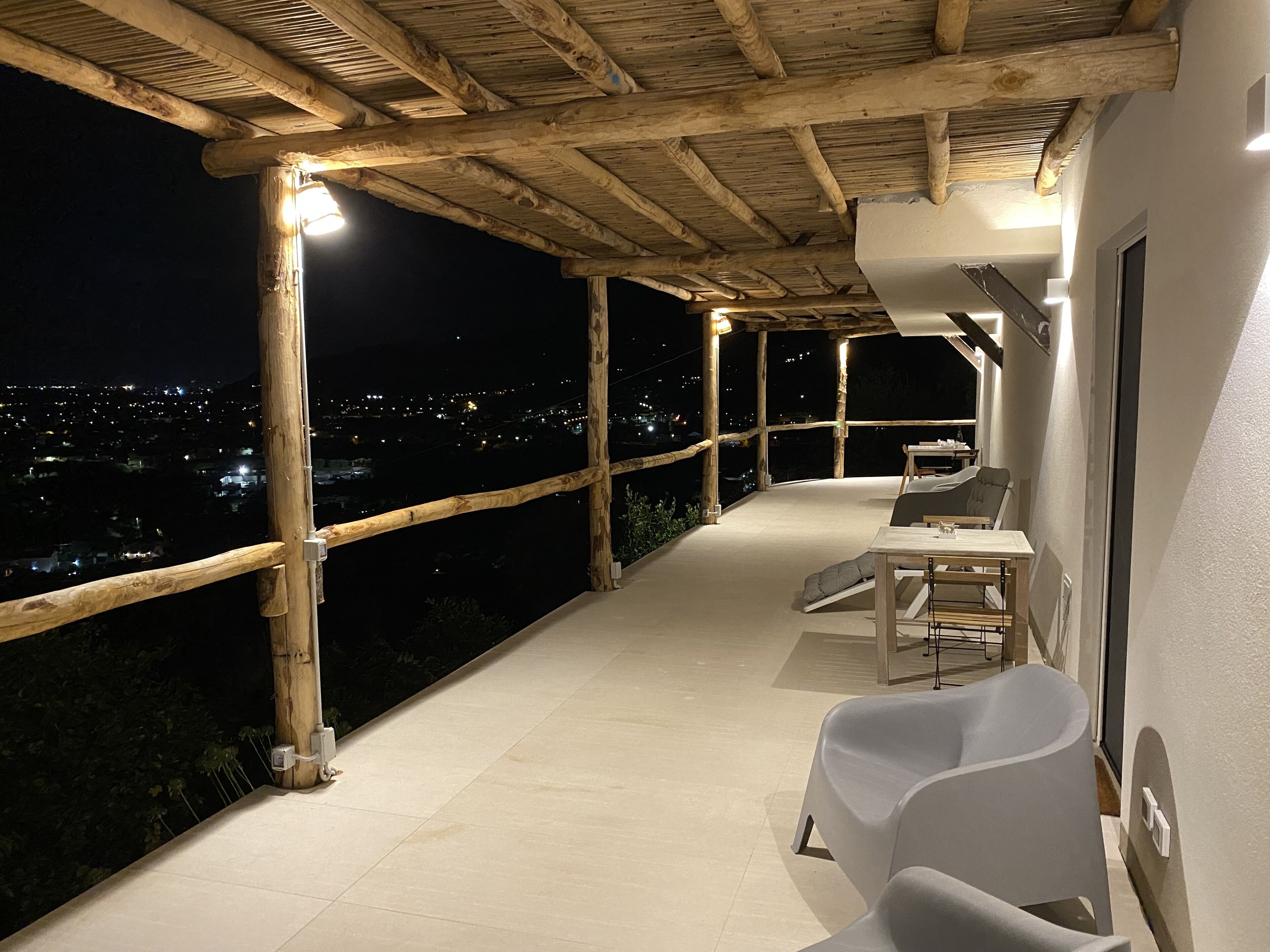 Una terraza techada al aire libre con zona de comedor, vigas de madera y vista a un paisaje urbano nocturno.