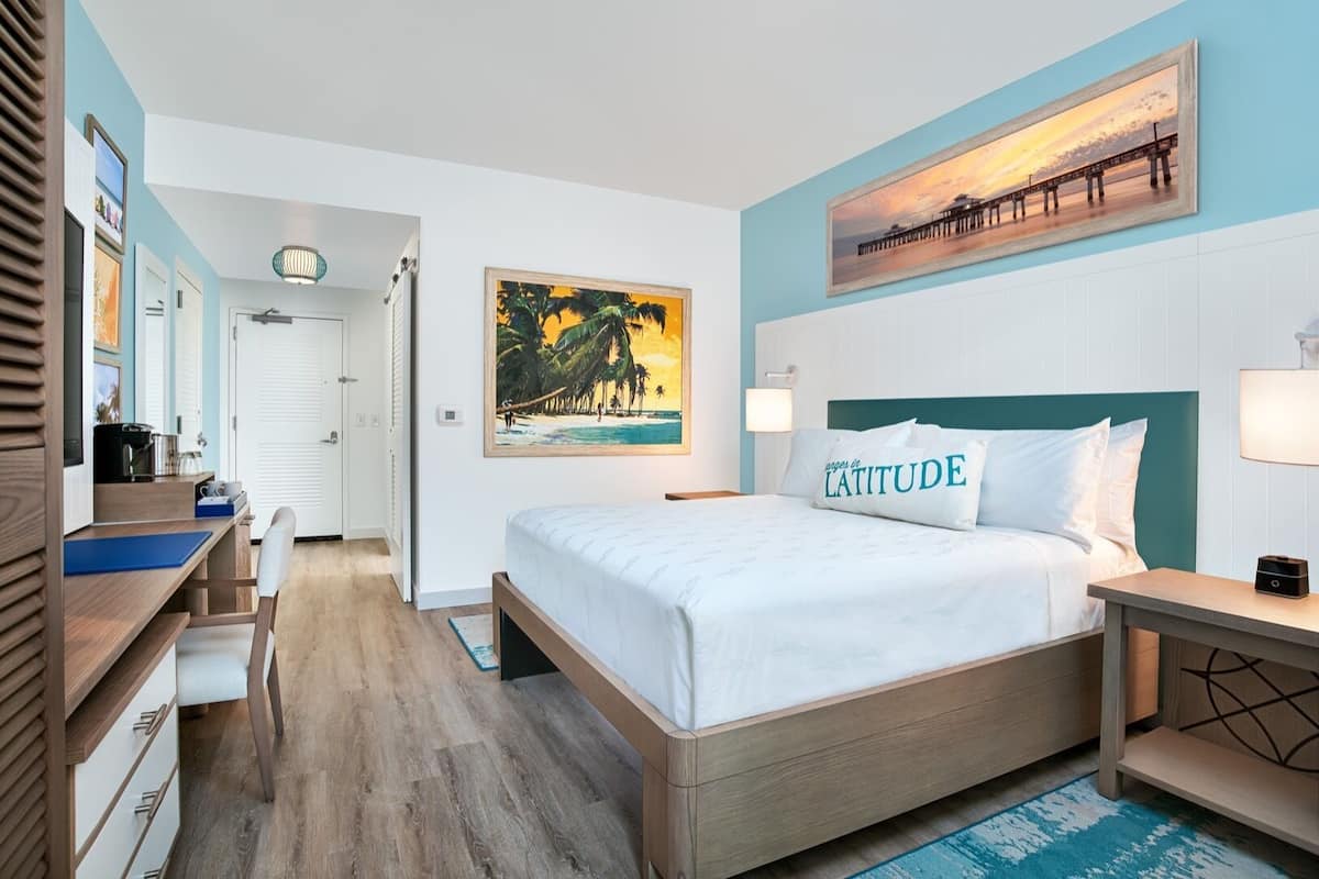 Une chambre d’hôtel avec un lit, un bureau, une chaise, une table de chevet et un tableau représentant un coucher de soleil sur une jetée.