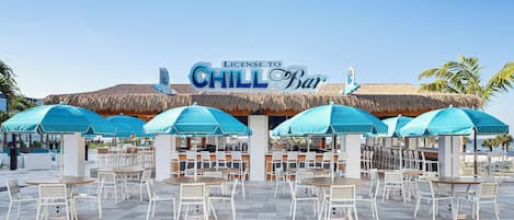 An outdoor seating area with tables and chairs, a thatched roof bar, and a sign reading "License to Chill Bar".