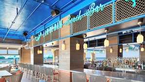 Un bar à thème plage, avec des meubles en osier, un comptoir bien garni de bouteilles variées et une vue sur la plage grâce à de grandes fenêtres.