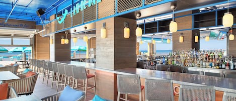 A beach-themed bar with wicker furniture, a bar counter with a variety of bottles, and a view of the beach through large windows.