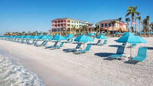 Une plage bordée de rangées de parasols bleus et de chaises longues, un immeuble doté de nombreux balcons et des palmiers.