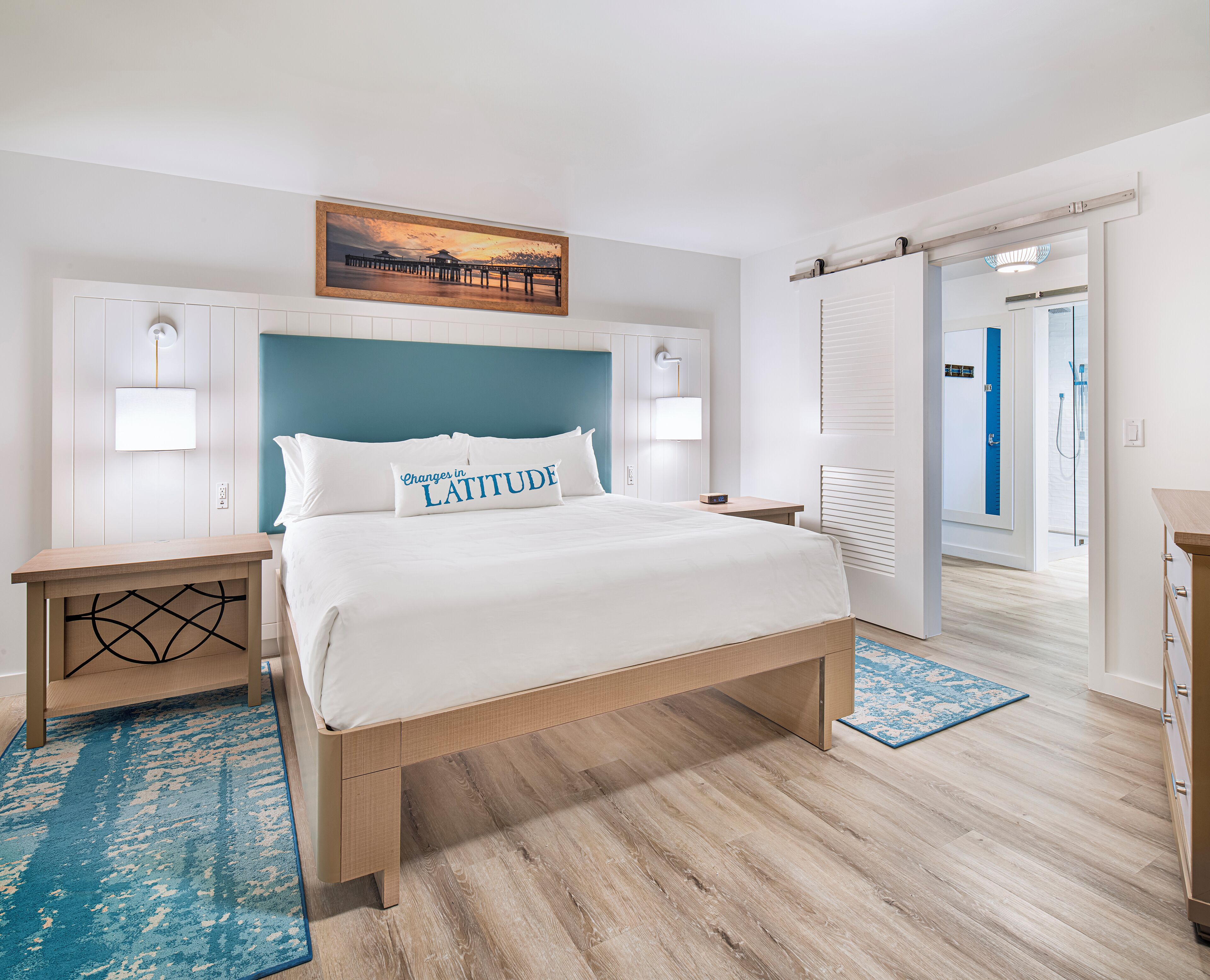 Een moderne hotelkamer met een groot bed, een nachtkastje, een wandlamp, een schilderij van een pier, en een badkamer die door een geopende deur zichtbaar is.