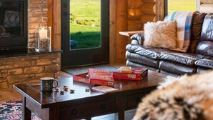 A cozy living room with a stone fireplace, leather sofa, and a view of a green field. - Luxe Log Cabin: Teton Views, Huge Patio, & Hot Tub (Tetonia)