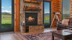 A log cabin interior with a stone fireplace, leather sofa, and a view of a grassy field and mountains. - Luxe Log Cabin: Teton Views, Huge Patio, & Hot Tub (Tetonia)