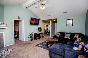A living room with a dark grey sofa, a ceiling fan, a TV mounted on the wall, and a fireplace. - Caleb’s King & Queen Home. 
Kids/Pet friendly, Family games, Piano. (Lubbock)