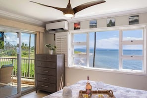 A room with a large window offering a view of the ocean, a ceiling fan, a dresser, and a table with a bottle and glasses.