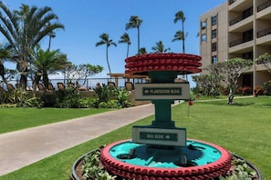 A fountain with a circular base and a top tier of red circular elements, surrounded by a green border. There are signs indicating directions to Plantation Bldg. and Mill Bldg. - Updated Oceanfront 1 bedroom condo, AC, 6 mile sandy beach, Central location (Kihei)