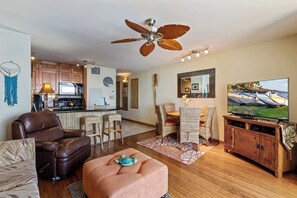 A living room with a sofa, ottoman, TV, and a dining area with chairs and a table. - Updated Oceanfront 1 bedroom condo, AC, 6 mile sandy beach, Central location (Kihei)
