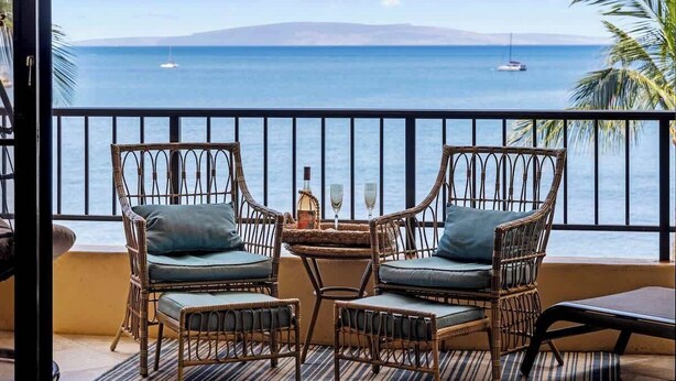 Un balcon agrémenté de meubles en osier, une table avec une bouteille et des verres, et une vue sur la mer avec des bateaux.