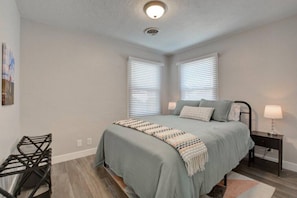 A bedroom with a bed, two bedside tables, a wall-mounted shelf, and a window with blinds.