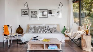 A modern living room with a bed, a coffee table, and a chair. There are framed pictures on the wall and a window with a view of trees.