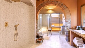 A modern bathroom with a mosaic tile wall, a shower with gold fixtures, a wooden bench, and a stone archway leading to a sauna area. - Villa Annabelle - Six Bedroom Villa, Sleeps 18 (Lucca)