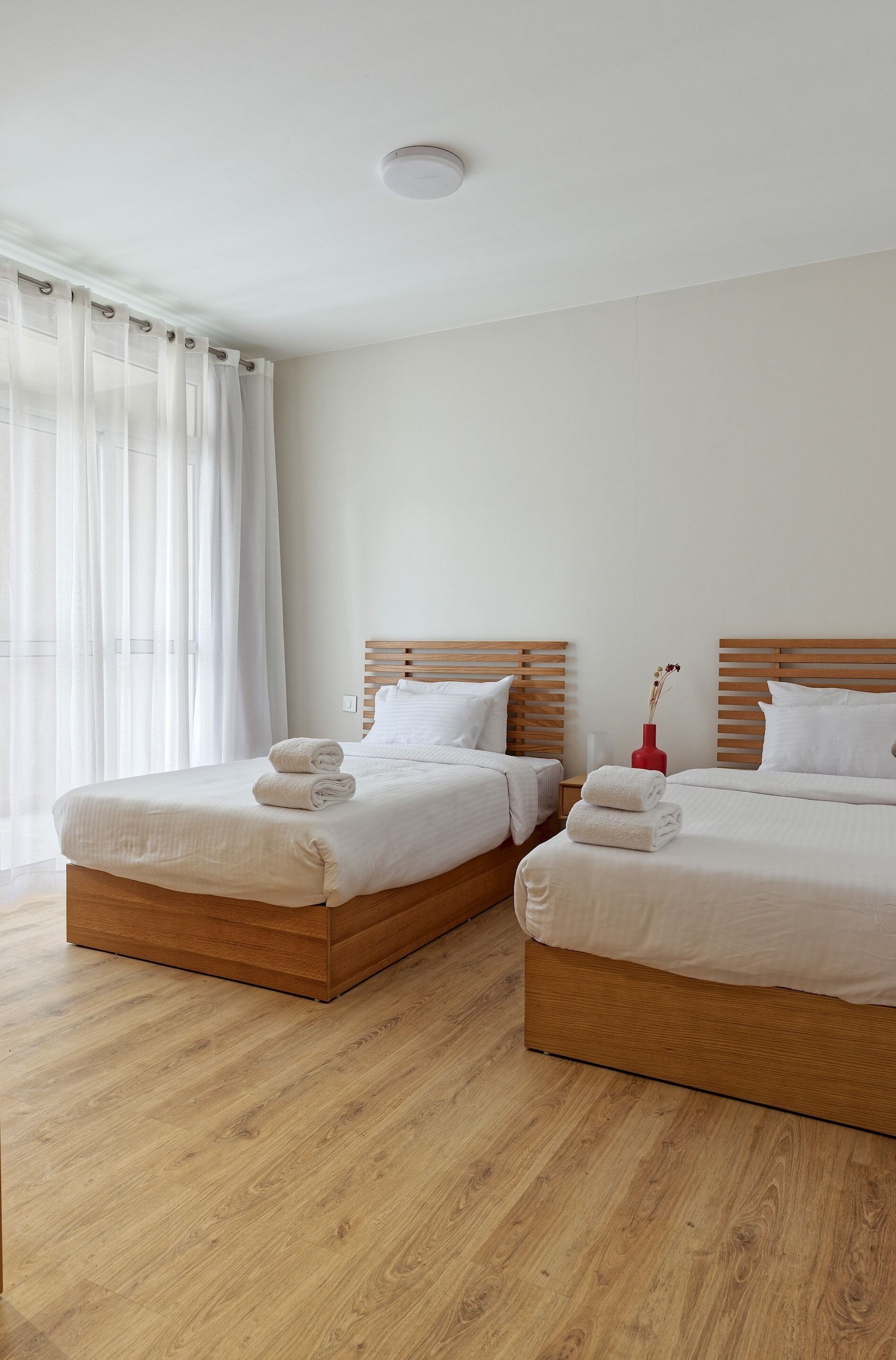 Habitación de hotel con dos camas, cabeceros de madera, ropa de cama blanca y un florero rojo sobre la mesita de noche.