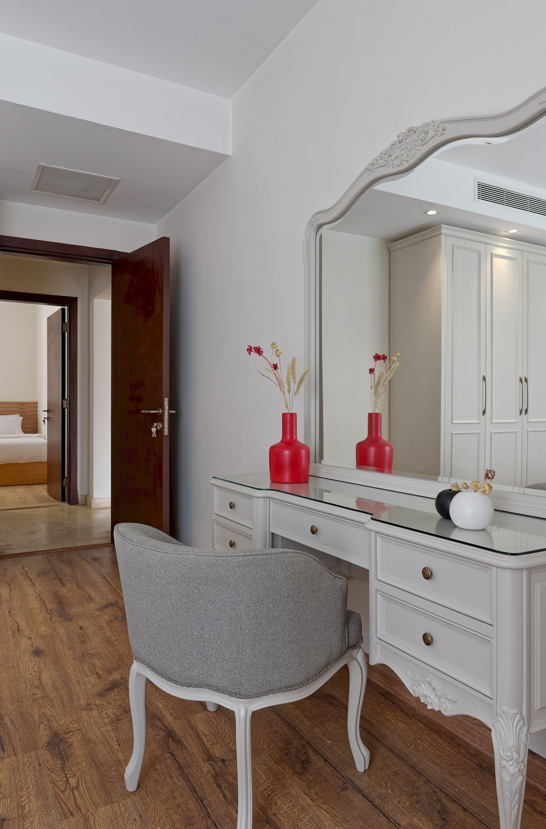 A room with a white vanity, a mirror, and a gray chair. There is a wooden door leading to another room.