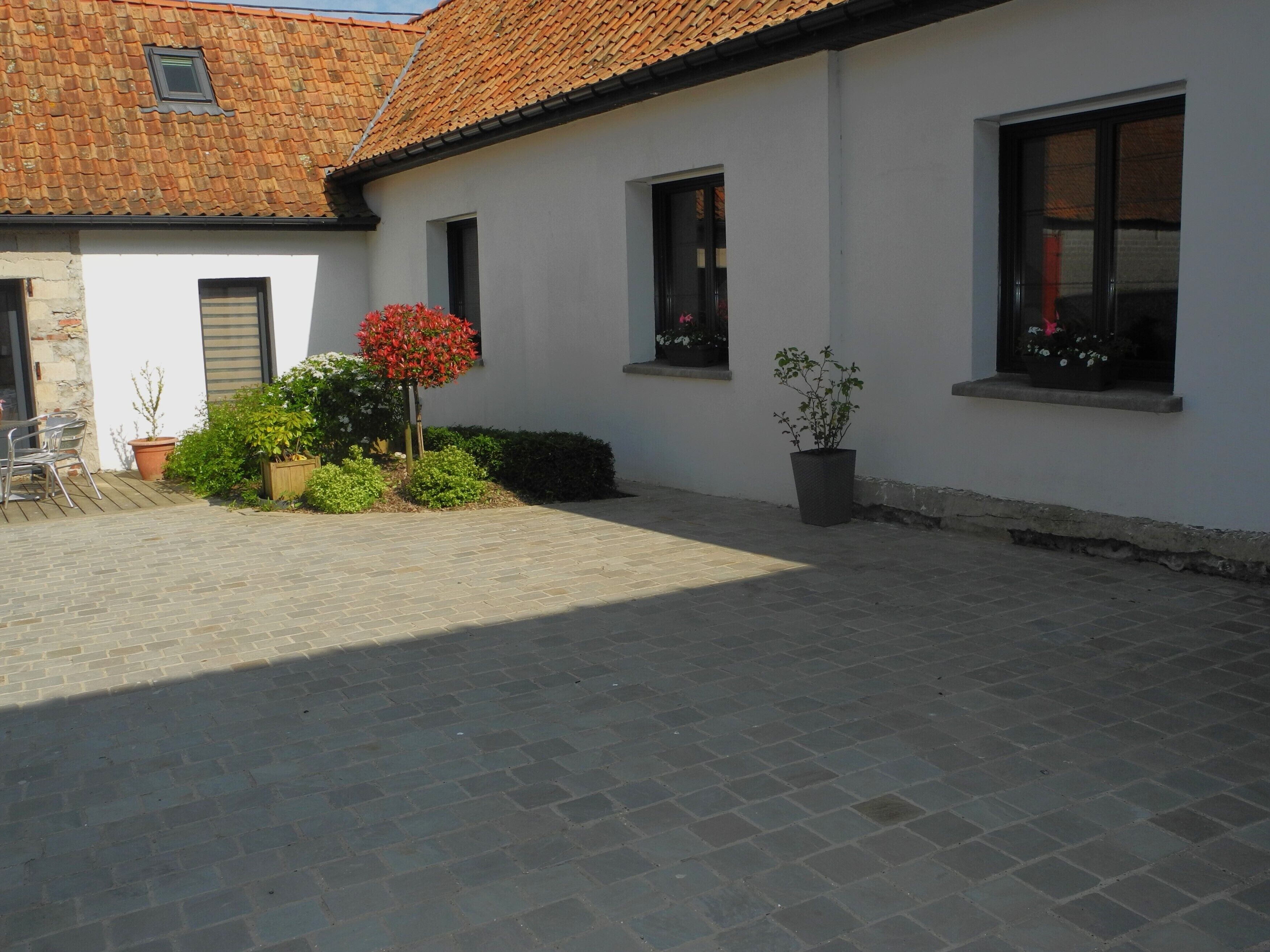 En innergård med en stenlagd gångväg, en vit byggnad med ett tak täckt av tegelpannor och krukväxter.