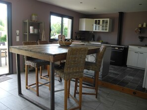 Un coin repas avec une table en bois et des chaises en osier, une cuisine avec des meubles blancs et une vue à travers une porte vitrée.