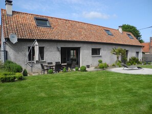 A house with a tiled roof, a satellite dish, a patio with a table and chairs, and a well-maintained lawn. - Gîte de Campagne (Widehem)