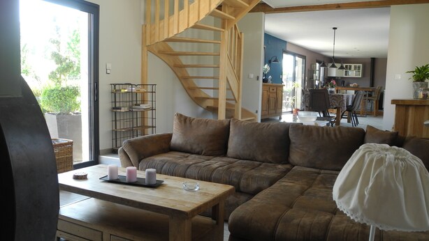 A cozy living room with a wooden staircase, a brown sofa, a wooden coffee table with candles, and a view of the kitchen in the background. - Gîte de Campagne (Widehem)