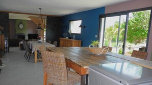 A dining area with a long wooden table and wicker chairs, a kitchen with a stove, and a large window with a view of greenery. - Gîte de Campagne (Widehem)