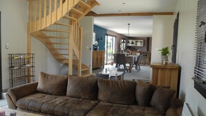 A living room with a brown leather sofa, a wooden staircase, a dining area with a table and chairs, and a kitchen in the background. - Gîte de Campagne (Widehem)