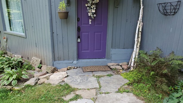 A house with a purple door, a stone pathway leading to the entrance, and a small garden area with plants and rocks. - BIRCH HAVEN (Bancroft)