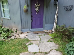 Une maison avec une porte violette, un chemin de pierres menant à l’entrée et un petit jardin agrémenté de plantes et de rochers.