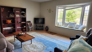 A living room with a sofa, a coffee table, a bookshelf, a TV, and a window with a view of greenery. - BIRCH HAVEN (Bancroft)