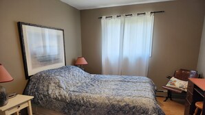 A bedroom with a bed, a desk, a chair, a lamp, a window with curtains, and a framed picture on the wall. - BIRCH HAVEN (Bancroft)