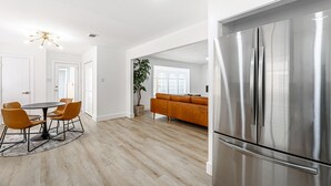 Una cocina moderna con un refrigerador de acero inoxidable, un comedor con una mesa redonda y sillas, y un espacio amplio y luminoso con un gran ventanal y una planta.
