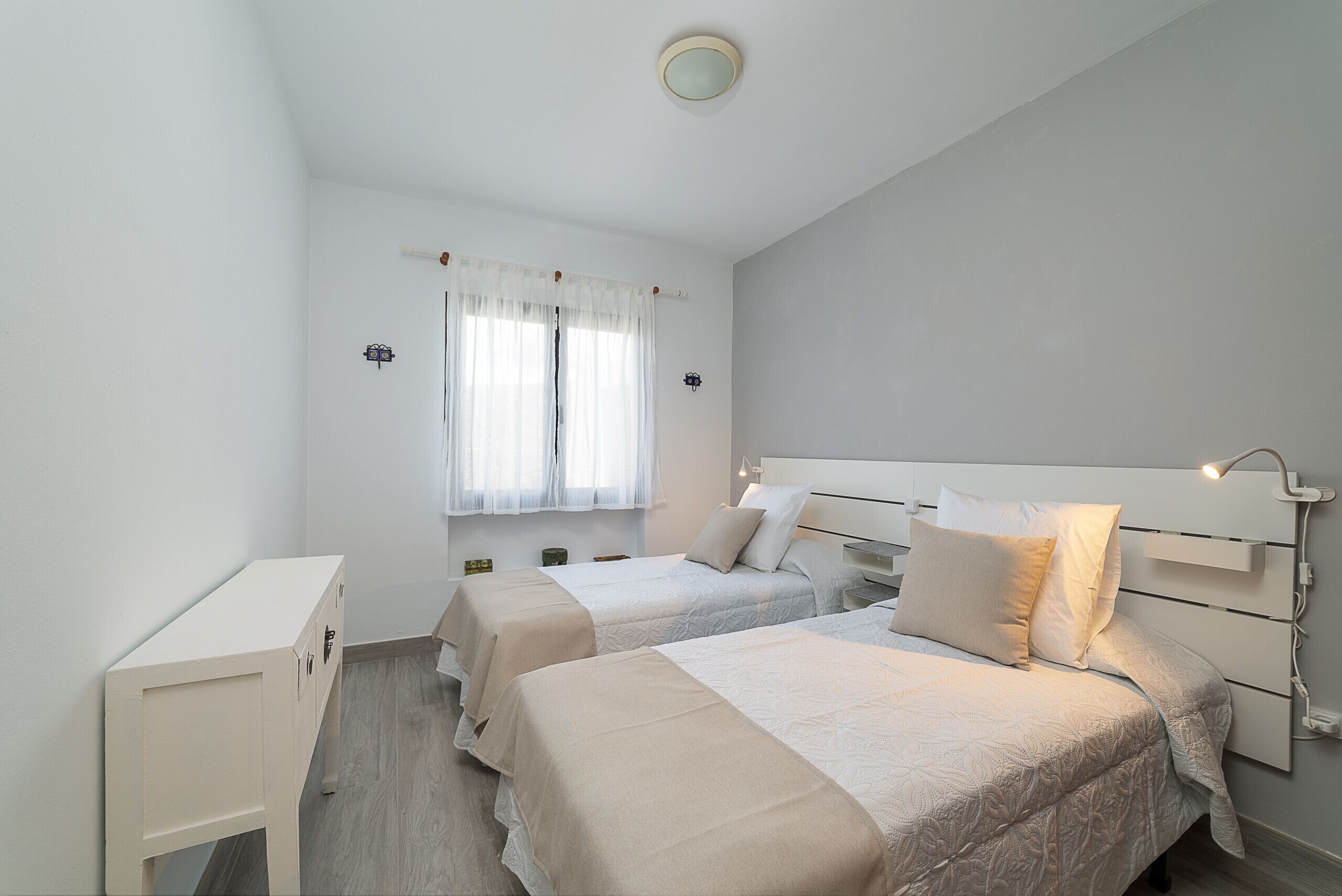 A bedroom with two beds, a bedside table, a nightstand, a window with curtains, and a ceiling light.