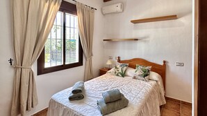 A bedroom with a bed, curtains, a nightstand, a lamp, and a window with a view of greenery.
