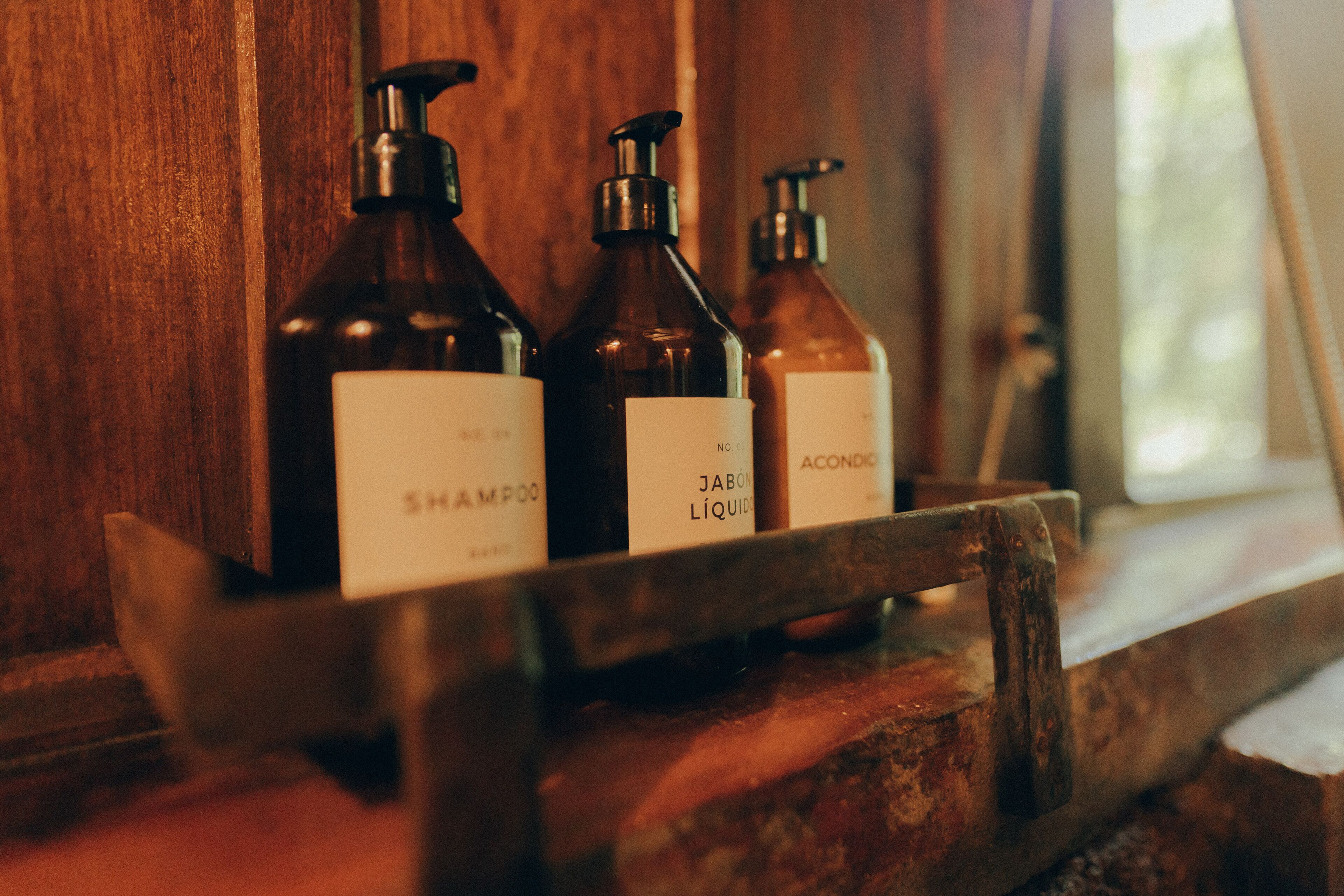 Three bottles of shampoo, a bottle of liquid soap, and a bottle of hand sanitizer on a wooden shelf.